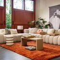 Moderne woonkamer met crème hoekbank, oranje vloerkleed, salontafel en sierkussens.