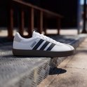 Witte Adidas sneaker met zwarte strepen, bruine zool, op de grond.