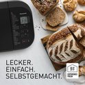 Zilverkleurige broodbakmachine met verschillende soorten brood, waaronder een met zaden erop.