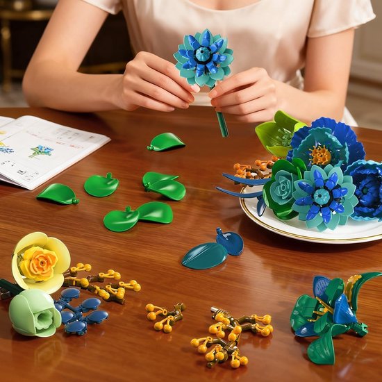 Een persoon bouwt een blauwe bloem met losse onderdelen op tafel. Er liggen meer bloemen en bladeren.