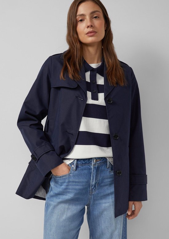 Model in donkerblauwe trenchcoat met ceintuur, gestreept shirt met brede blauwe banden, en wijde jeans.