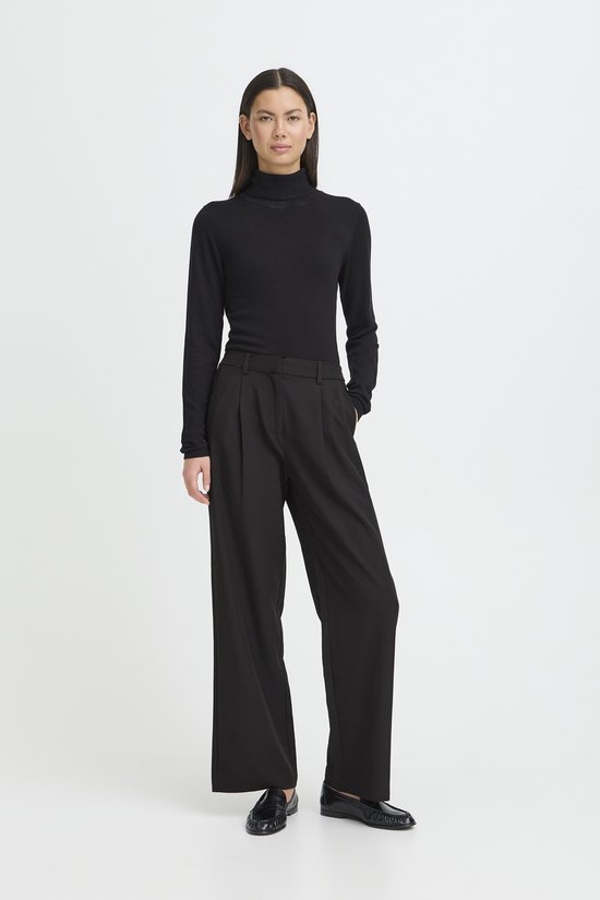 Een vrouw draagt een zwarte coltrui en een zwarte wijde broek met plooien.