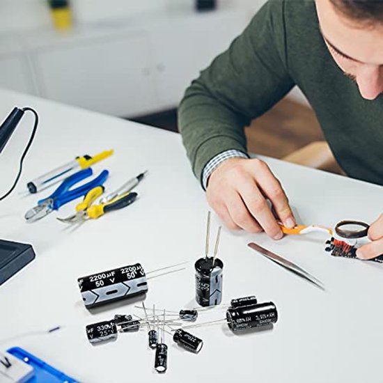 Diverse elektrolytische condensatoren op een witte tafel, naast gereedschap en een persoon die soldeert.