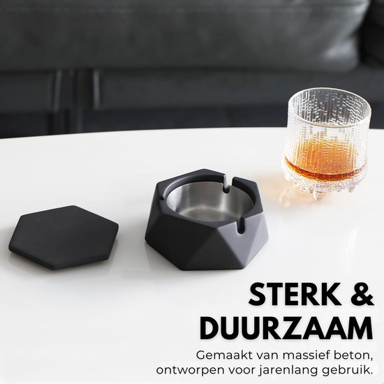 Zwarte, zeshoekige asbak van massief beton op witte tafel, met deksel ernaast en een glas whisky.