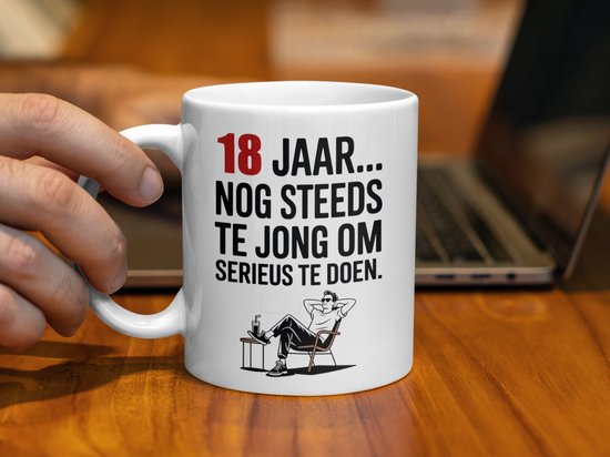 Witte mok met tekst "18 JAAR... NOG STEEDS TE JONG OM SERIEUS TE DOEN." en illustratie van een relaxte man in een stoel.