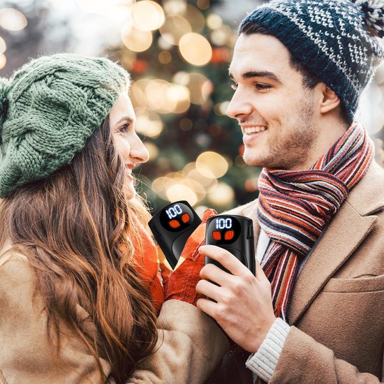 Koopel in winterkleding houdt zwarte handwarmers met digitaal display "100" vast. Kerstboom op achtergrond.