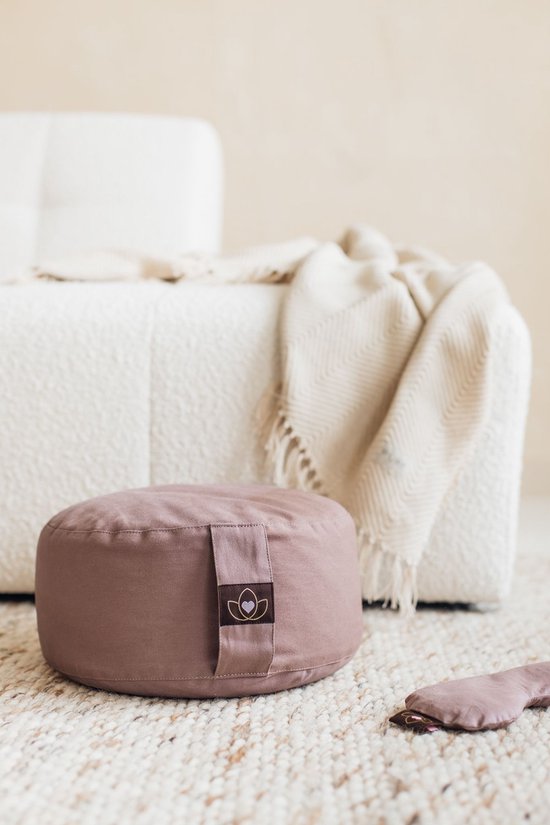 Taupe meditatiekussen op een vloerkleed, naast een bank en een oogmasker.
