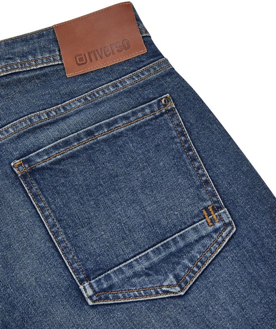 Detail van de achterkant van een blauwe jeans met twee zakken en bruin leren logo 'riverso'.