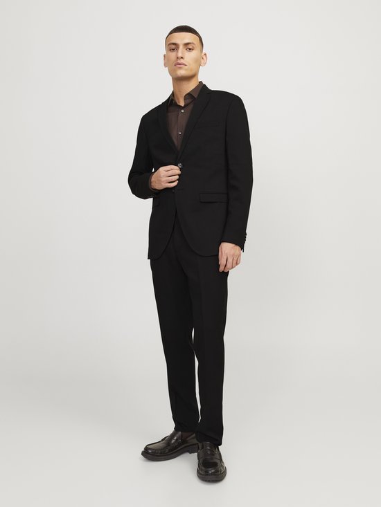 Man in zwart JACK & JONES blazer en broek met bruine blouse en bruine instappers. Hij poseert met de hand op de blazer.