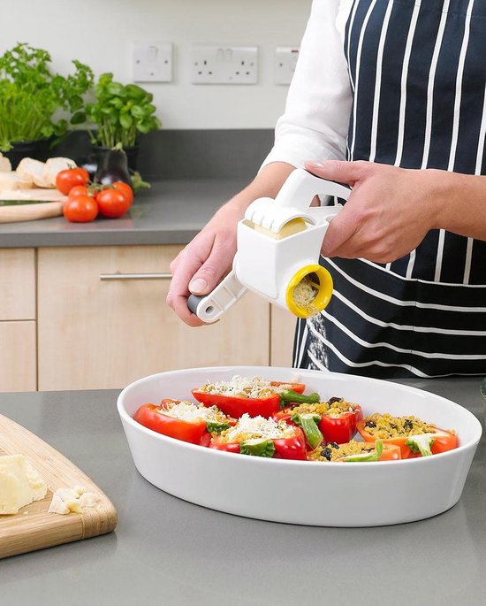 Persoon met gestreept schort gebruikt een witte kaasrasp met gele opening om Parmezaanse kaas over gevulde pepers te raspen.