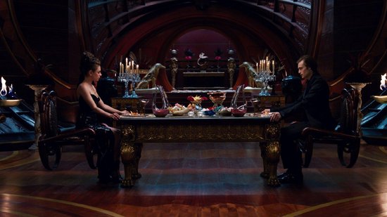 Een man en vrouw zitten tegenover elkaar aan een grote, versierde tafel in een weelderige kamer.