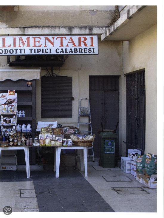 Buitenwinkel met een uithangbord met 'ALIMENTARI ODOTTI TIPICI CALABRESI'. Tafels met etenswaren en een ladder.