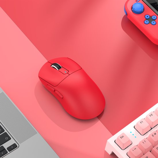 Een rode gaming muis op een bureau naast een laptop en een roze toetsenbord.