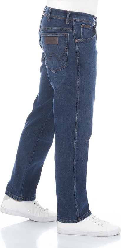Zij-aanzicht van een man in een blauwe Wrangler jeans met bruine leren details en witte sneakers.
