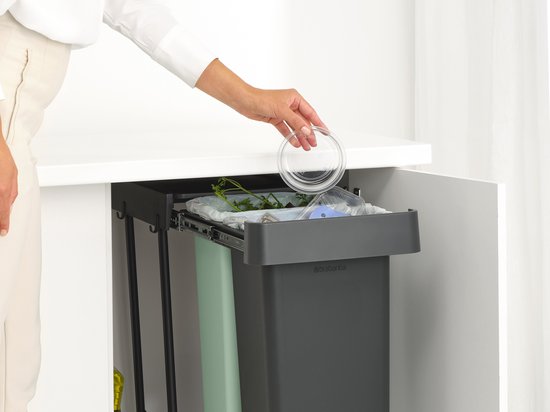 Een hand die een deksel in een donkergrijze Brabantia prullenbak legt, in een keukenkastje.