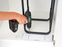 Een hand gebruikt een boormachine om een zwart metalen frame voor prullenbakken te monteren.
