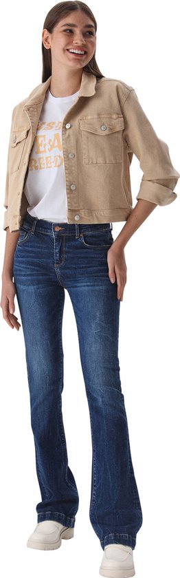 Model van voren in een donkerblauwe flared spijkerbroek, beige jack en witte sneakers.