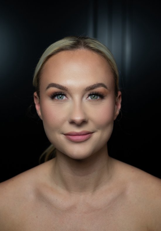 Model met blond haar, groene ogen, en subtiele make-up, lachend naar de camera.