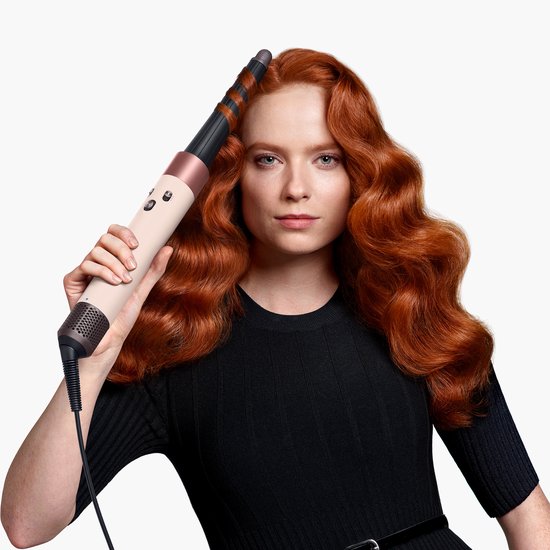 Vrouw met rood golvend haar houdt een beige en rosé-gouden Dyson Airwrap omhoog.