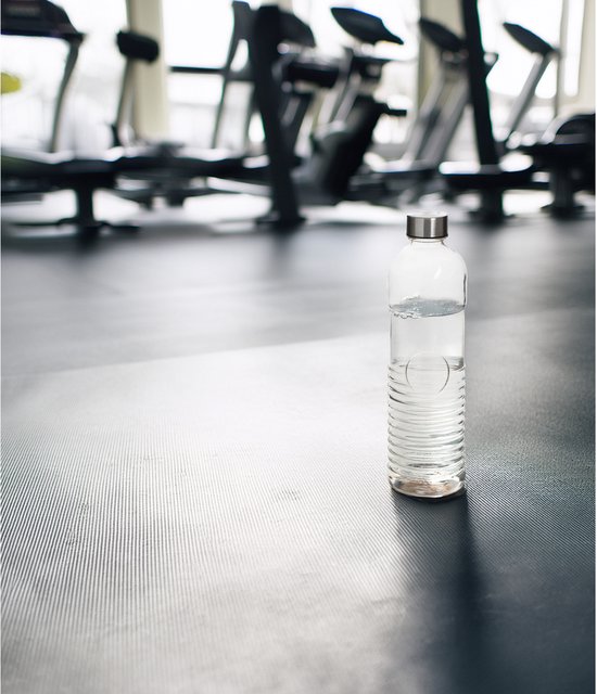 Glazen waterfles van 1 liter op de vloer van een fitnessruimte, naast loopbanden.