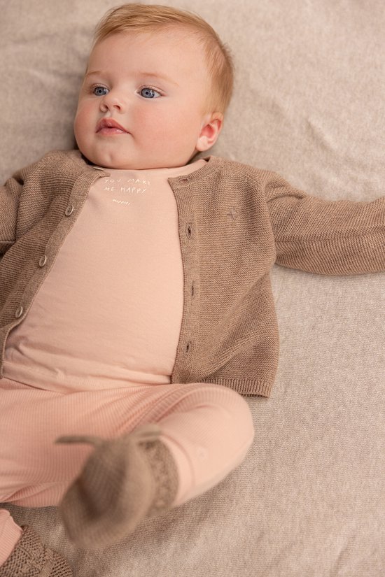 Baby met blauwe ogen, draagt een roze longsleeve shirt onder een bruin vest en een roze broek.