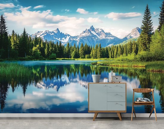 Paysage de montagne avec sommets enneigés, pins, lac et son reflet, avec des meubles : une commode bleue et une chaise en bois.