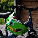 Lichtgroene kinderfietshelm in dinosaurusvorm, met blauwe ogen en scherpe tanden. Hangt aan een fietsstuur.