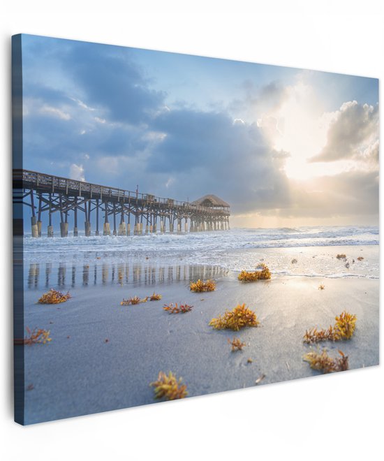 Canvas van een pier aan zee met reflecties en zeewier op het zand bij zonsondergang.
