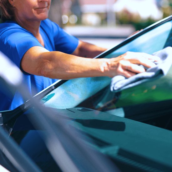 Une femme en chemise bleue nettoie la vitre d'une voiture avec un chiffon gris.
