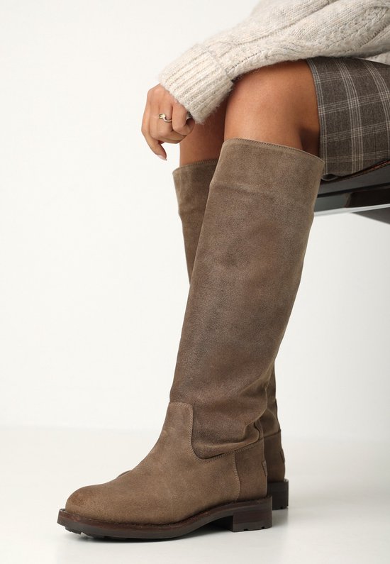 Model zittend op een stoel, draagt taupe suède kniehoge laarzen, een geruite rok en een gebreide trui.