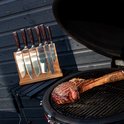 Set van zes damascus messen met donkerhouten handvatten op houten messenhouder, met een groot stuk vlees op de grill.
