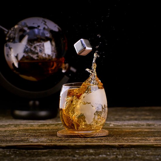 Een glas met amberkleurige drank en een wereldkaartmotief, met een splash en een metalen ijsblokje dat eruit springt. Een whiskybolvormig glas staat op de achtergrond.