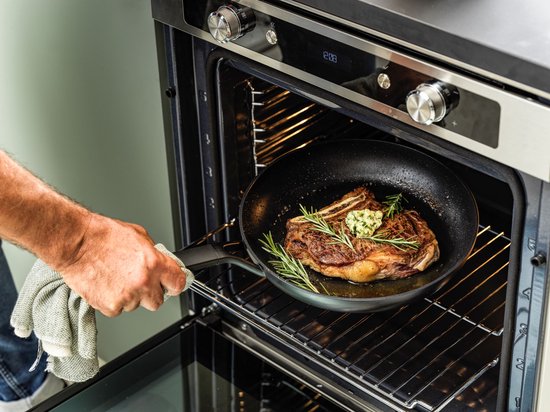 Man plaatst koekenpan met grote biefstuk en rozemarijn in een oven.