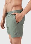 Homme avec la main dans la poche d'un short de bain vert vintage avec logo noir sur la jambe gauche.