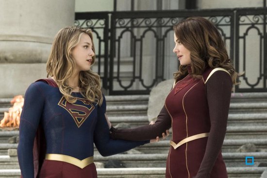 Supergirl met blauw pak en rood masker staat naast een vrouw met rood pak.