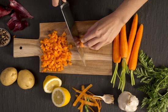 Bovenaanzicht: chef snijdt wortelen in blokjes op houten plank met Tefal mes. Citroenen, aardappelen, knoflook, ui, peperbolletjes erbij.