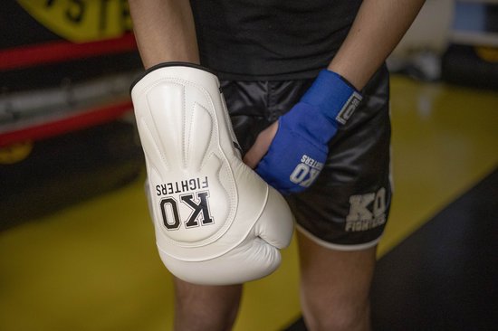 Een persoon met blauwe bandages om de linkerhand en een witte KO FIGHTERS bokshandschoen om de rechterhand.