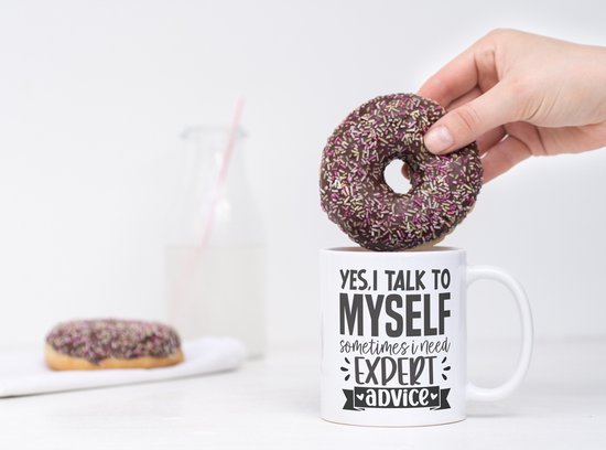 Witte mok met tekst "YES, I TALK TO MYSELF SOMETIMES I NEED EXPERT ADVICE", met een donut die erin wordt gestoken.