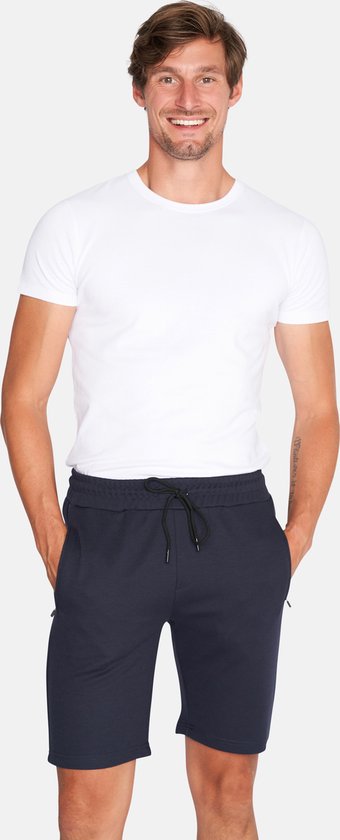 Man met witte t-shirt en donkerblauwe korte broek met trekkoord, staand en lachend.