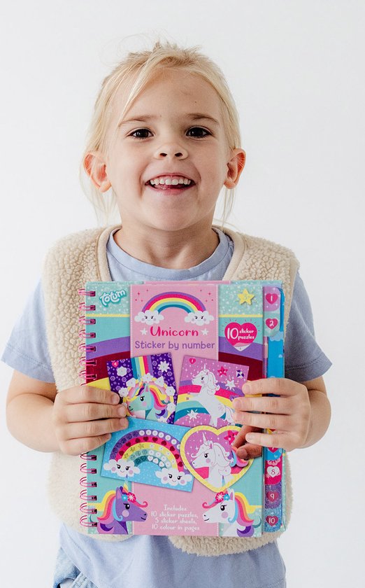 Une jeune fille joyeuse aux cheveux blonds et portant un gilet crème tient un livre "Sticker by number" Licorne. Le livre présente un arc-en-ciel, des nuages et des licornes.