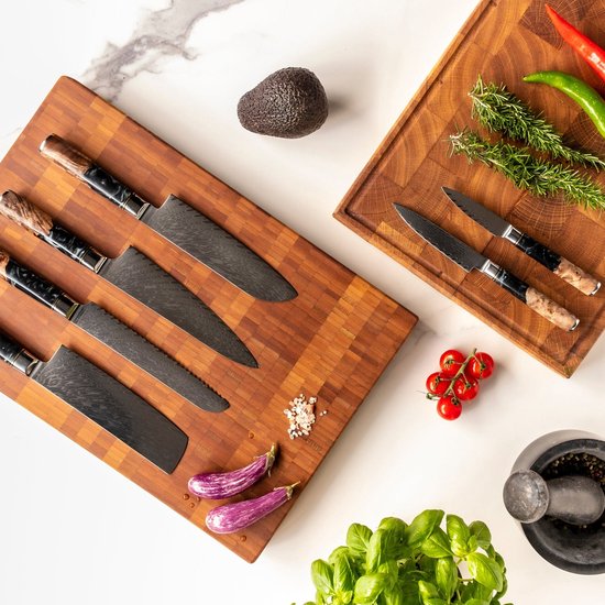 Een set van vijf Japanse messen met damastpatroon en unieke handvatten van hout en blauwe epoxy, op een houten snijplank met avocado, tomaten, aubergines en kruiden.