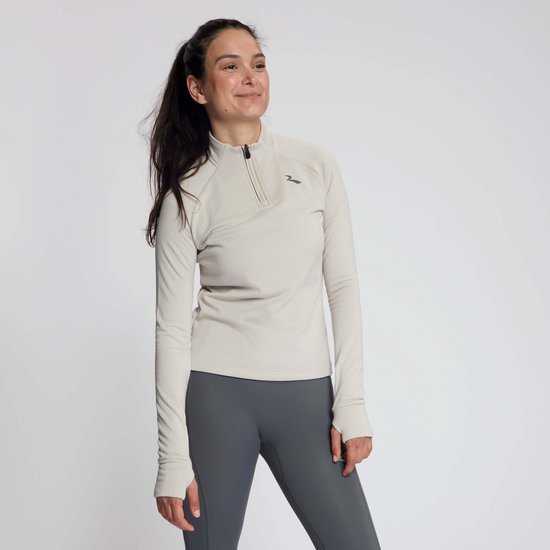 Een vrouw draagt een zandkleurig sportshirt met halve rits en grijze legging. Ze lacht en kijkt opzij.