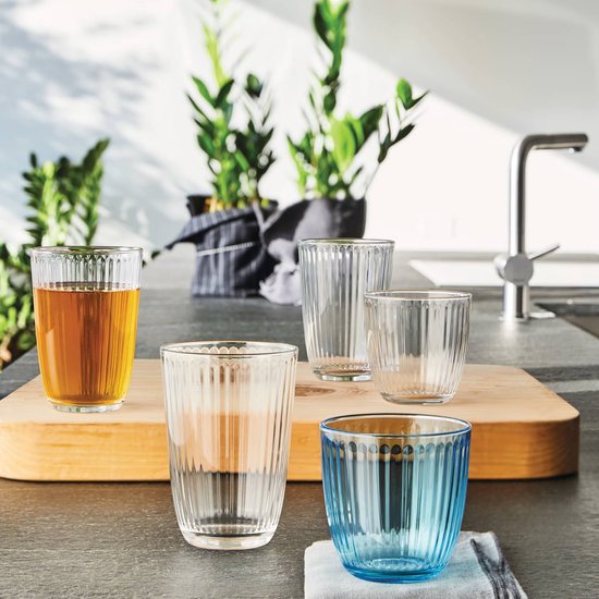 Plusieurs verres tumbler côtelés sur une planche en bois, avec une plante et un robinet à l'arrière.
