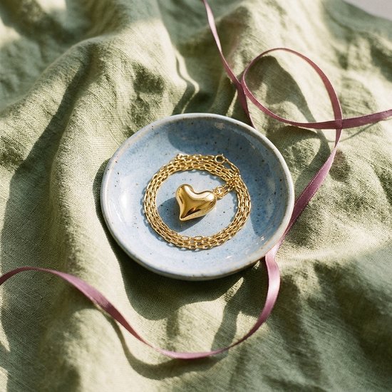 Goudkleurige ketting met hartvormige hanger in een lichtblauw schaaltje op een groene stoffen achtergrond met lint.
