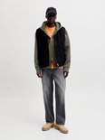Model draagt een zwart gilet over een olijfgroene hoodie, oranje shirt, grijze jeans en beige schoenen.