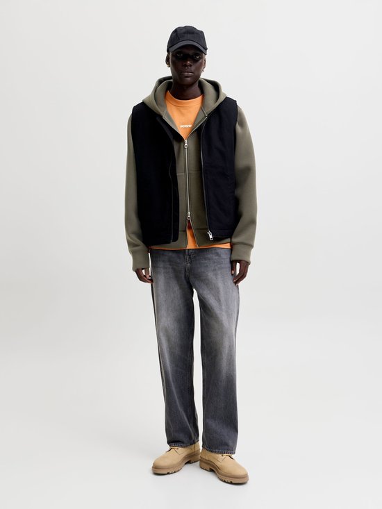 Model draagt een zwart gilet over een olijfgroene hoodie, oranje shirt, grijze jeans en beige schoenen.