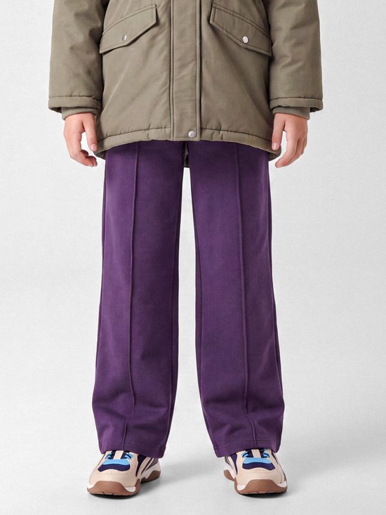 Close-up van de paarse joggingbroek met wijde pijpen gedragen onder een kaki jas, toont de persvouw op de pijpen.