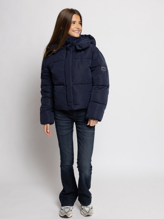 Meisje met lang bruin haar in een donkerblauwe pufferjas, spijkerbroek en sneakers, lachend naar de zijkant.