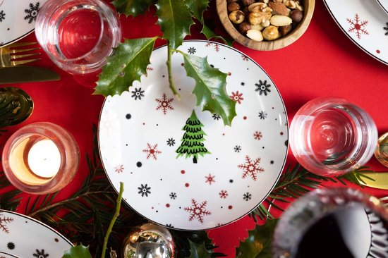 Feestelijk gedekt wit bord met kerstdecoratie, midden op een rode tafelkleed omringd door hulst.