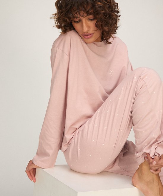 Model in een zachtroze pyjamaset met lange mouwen en een subtiel hartje op de borst. De pyjamabroek is bedekt met kleine witte hartjes.